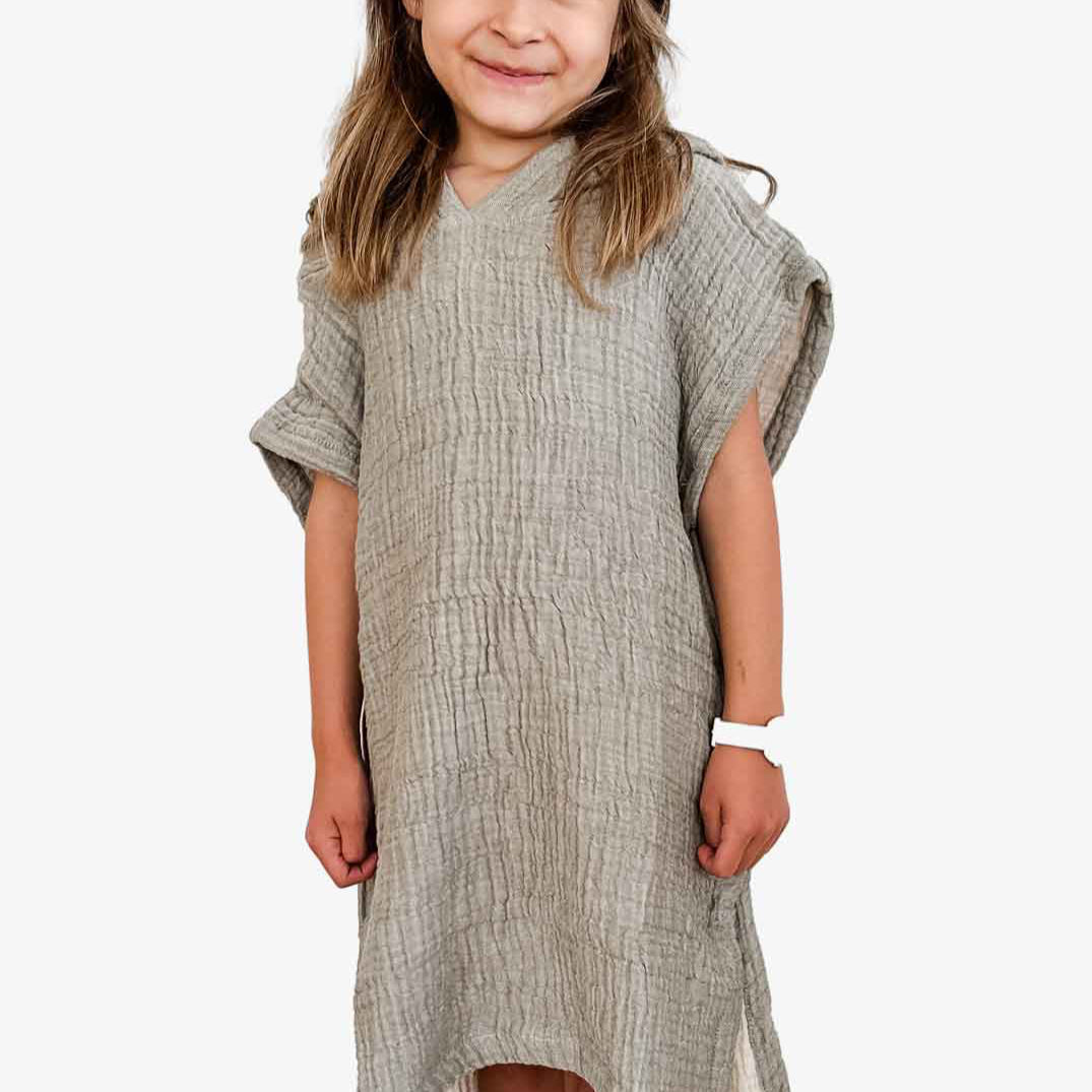 Ein junges Mädchen mit langen Haaren lächelt in die Kamera. Es trägt einen locker sitzenden, strukturierten olive grünen Kinder Badeponcho Flow von Main Design aus OEKO-TEX zertifizierter Baumwolle. Sie trägt ein weißes Armband auf einem schlichten weißen Hintergrund, der ihren fröhlichen Ausdruck unterstreicht.