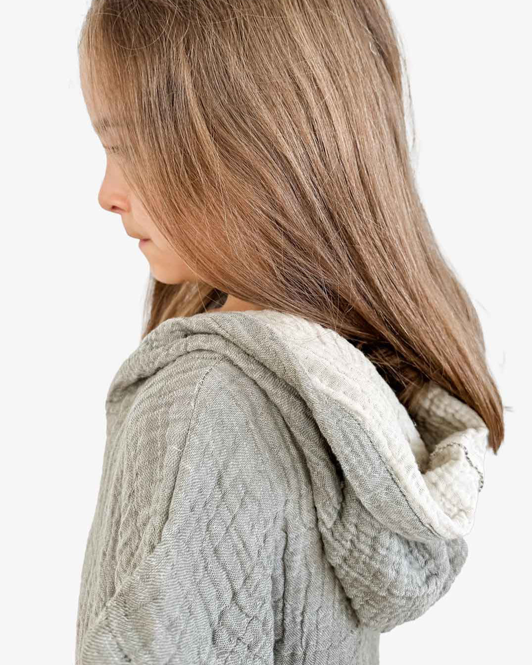 Ein Kind mit langen braunen Haaren ist im Profil abgebildet und trägt vor einem schlichten weißen Hintergrund einen strukturierten hellgrauen Kinder Badeponcho Flow von Main Design aus OEKO-TEX-zertifizierter Baumwolle.