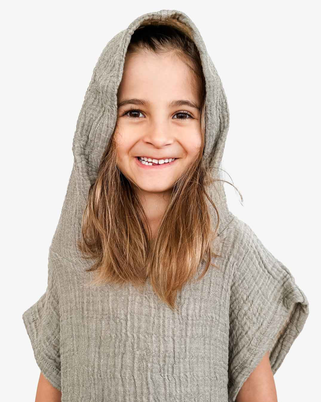Ein lächelndes Kind mit langen Haaren trägt einen strukturierten Kinderbadeponcho Flow mit Kapuze von Main Design, gefertigt aus OEKO-TEX zertifizierter Baumwolle. Der schlichte weiße Hintergrund betont den fröhlichen Ausdruck des Kindes und den weichen Musselinstoff.