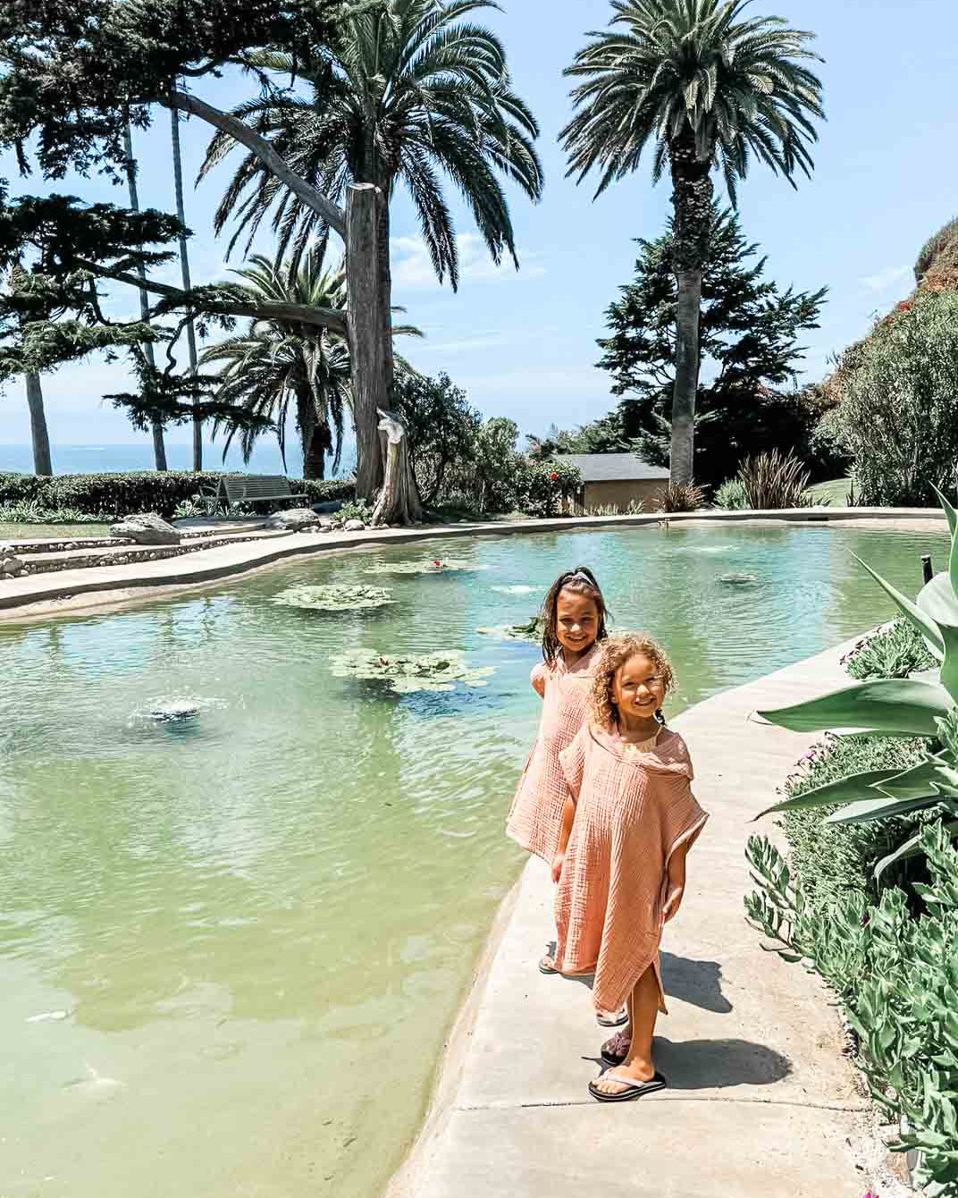 Zwei Kinder stehen an einem Pool und tragen passende hellrosa Kinder Badeponcho Flow-Outfits von Main Design, hergestellt aus OEKO-TEX zertifizierter Baumwolle. Der Pool liegt in einem üppigen Garten mit Palmen vor einem klaren blauen Himmel.