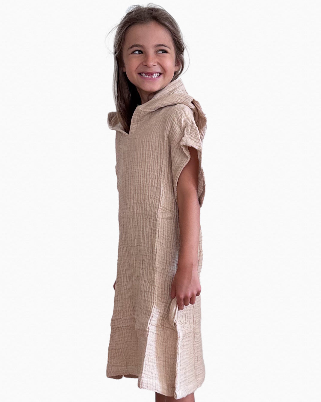 Ein lächelndes kleines Kind mit teilweise zurückgestecktem braunem Haar steht seitlich vor einem weißen Hintergrund und trägt den strukturierten beigen Main Design Kinder Badeponcho Flow aus OEKO-TEX zertifizierter Baumwolle mit kurzen Ärmeln.
