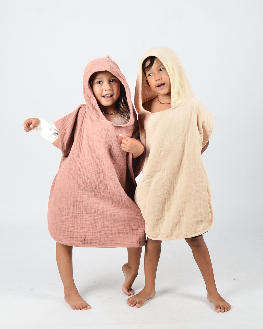 Zwei lächelnde Kinder stehen barfuß auf einem einfarbigen Hintergrund, gekleidet in den kuscheligen Main Design Kinder Badeponcho Flow. Das eine trägt Rosa, das andere Beige, aus weichem Musselin aus OEKO-TEX zertifizierter Baumwolle. Mit umeinander geschlungenen Armen nehmen sie eine verspielte Pose ein.