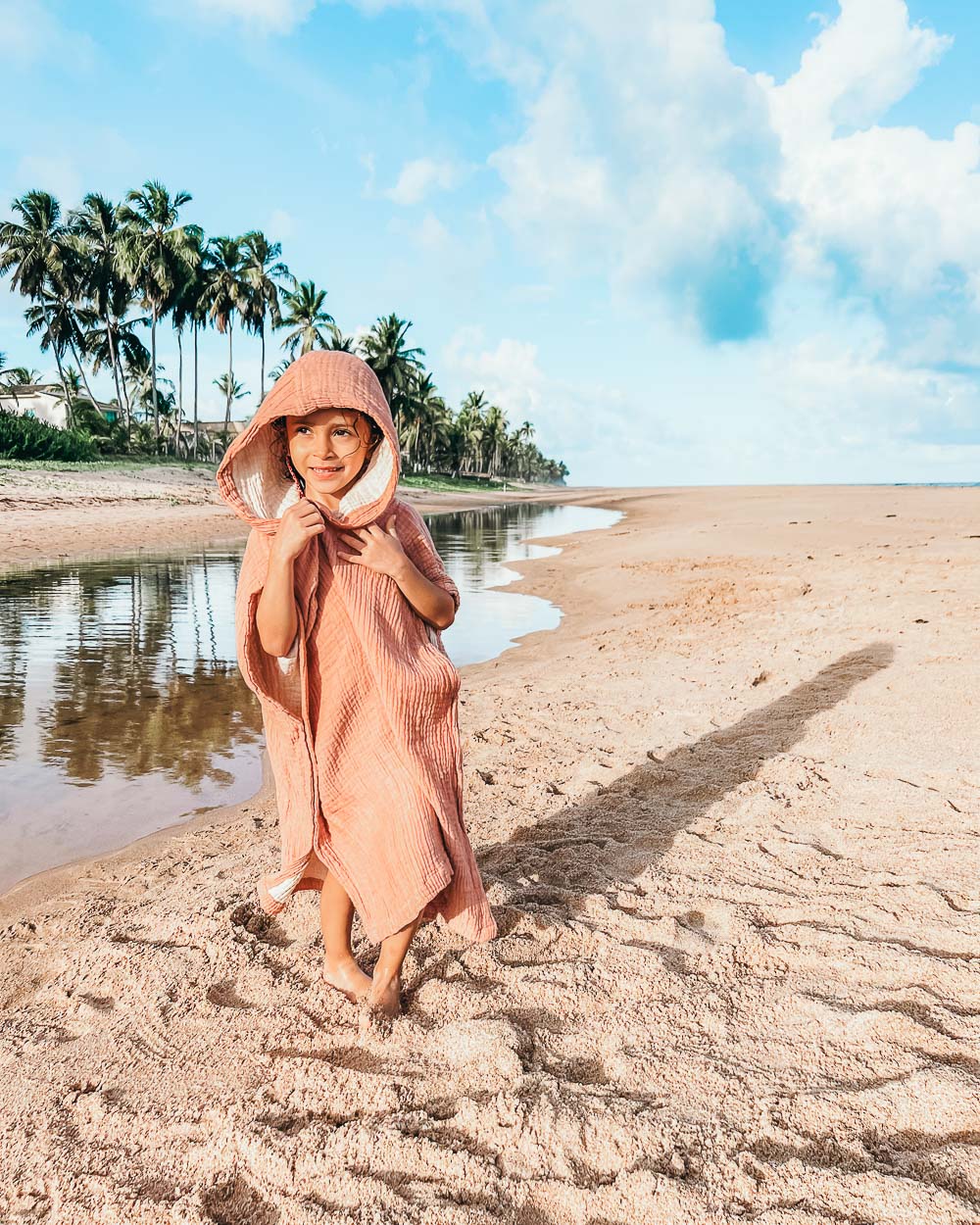 Ein Kind in einem rosa Main Design Kinder Badeponcho Flow aus OEKO-TEX Baumwolle steht barfuß und lächelnd an einem Sandstrand. Palmen, blauer Himmel und ein nahe gelegener Bach vervollständigen die Szene.