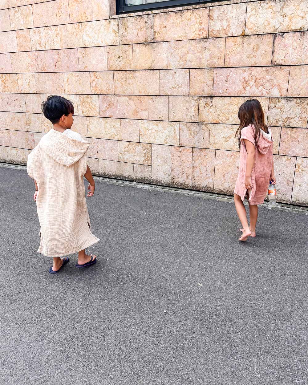Zwei Kinder im Main Design Kinder Badeponcho Flow laufen barfuß auf einem gepflasterten Weg an einer Steinmauer. Das eine trägt einen leichten Poncho mit Sandalen, das andere ein rosafarbenes Musselin-Handtuch und ist barfuß - beide schauen beim Gehen nach unten.