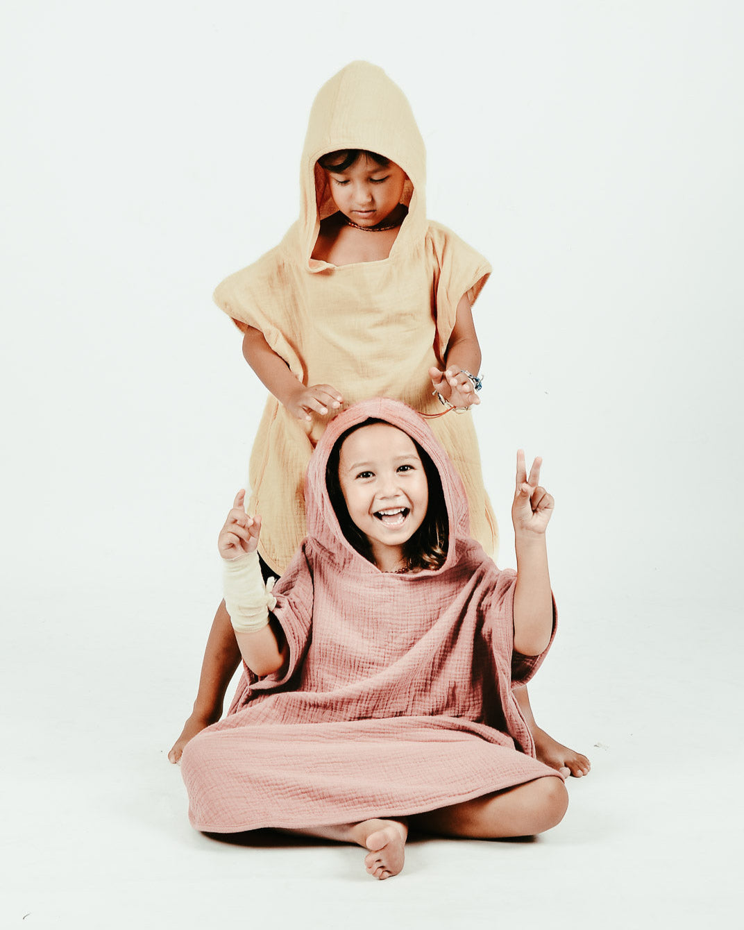 Zwei Kinder sitzen verspielt vor einem schlichten Hintergrund. Das vordere Kind, in einem rosa Kapuzenoutfit aus OEKO-TEX-zertifizierter Baumwolle, lächelt und zeigt Peace-Zeichen. Das hintere Kind trägt den Main Design Kinder Badeponcho Flow in Gelb und blickt mit Freude und Zufriedenheit im Gesicht nach unten.