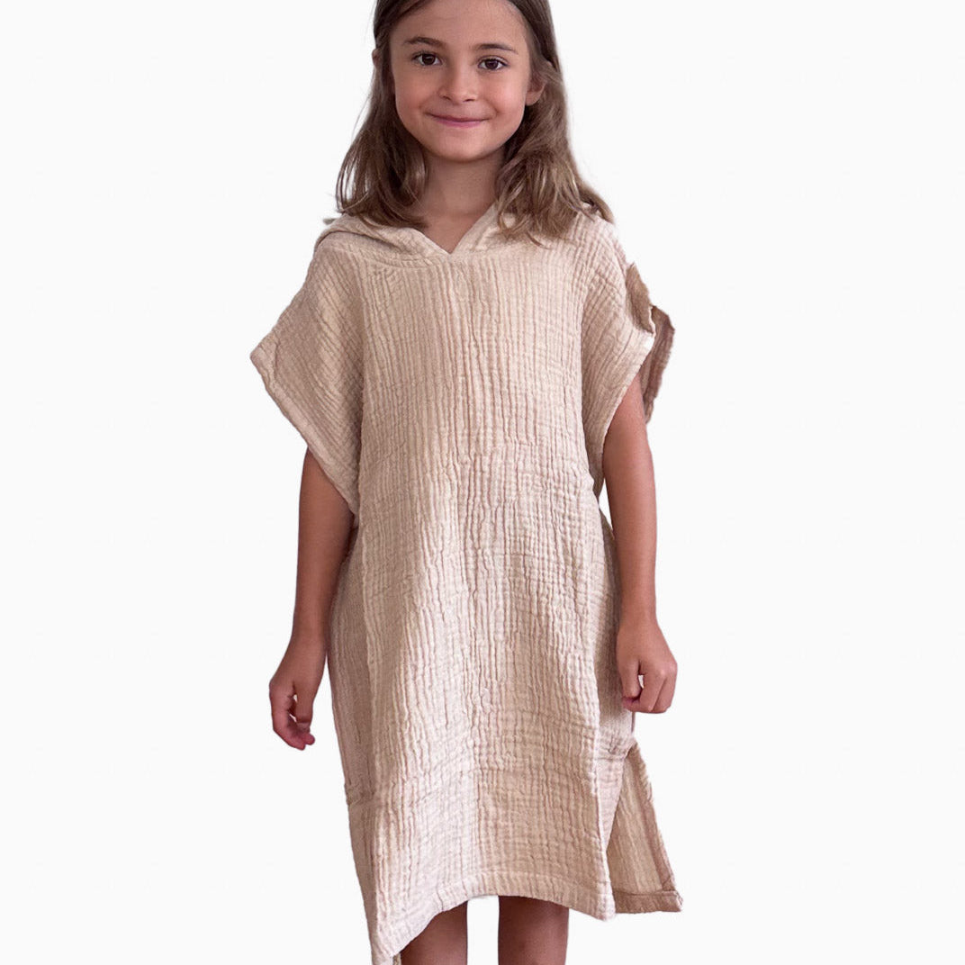 Ein junges Mädchen mit langen braunen Haaren steht barfuß und lächelt. Sie trägt ein locker sitzendes, strukturiertes beiges Kleid aus Musselin von Main Design aus OEKO-TEX-zertifizierter Baumwolle. Das Produkt ist der Kinder Badeponcho Flow, der vor einem schlichten weißen Hintergrund steht.