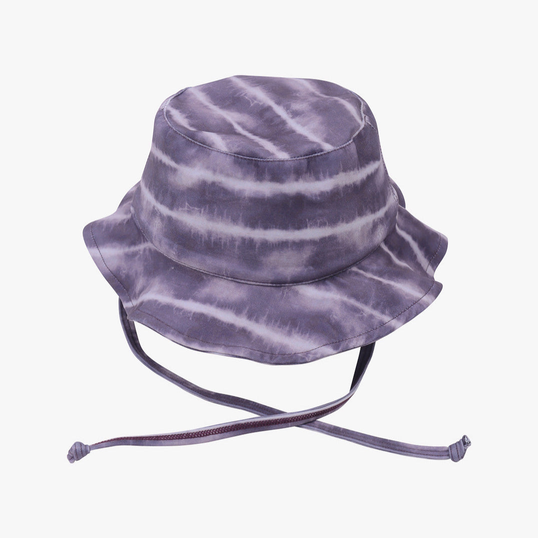 Ein lilafarbener **Kinder-Sonnenhut** mit Batikmuster, breiter Krempe und verstellbaren Kinnriemen, hergestellt aus umweltfreundlichem Stoff. Der Hut weist ein Muster aus horizontalen Streifen in verschiedenen Lila- und Weißtönen auf und bietet hervorragenden Sonnenschutz (LSF 50+). Er ist von **Main Design**.