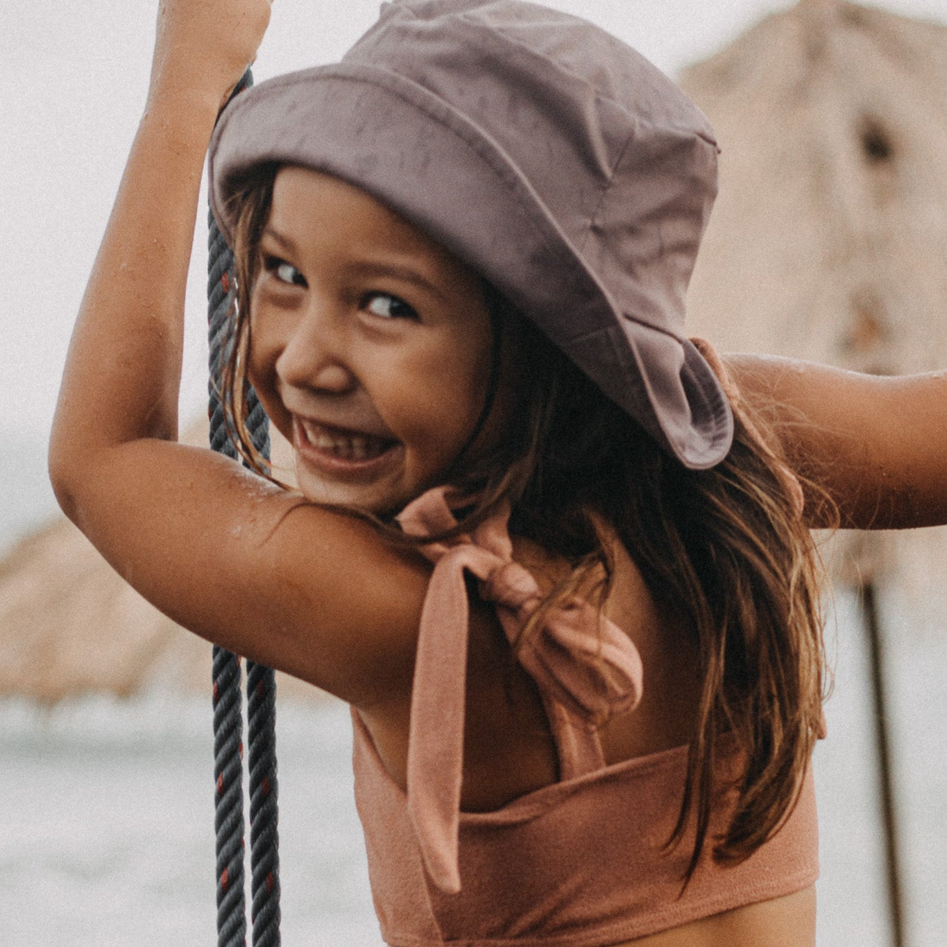 Ein junges Mädchen in einem hellbraunen Bikini und einem Kinder-Sonnenhut von Main Design lächelt und hält ein Seil. Sie steht an einem sonnigen Strand mit dem Meer und den Felsen im Hintergrund und genießt den Sonnenschutz UPF 50+.