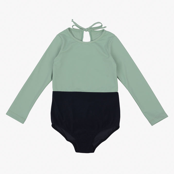Der Laila Mädchen Badeanzug von Main Design ist ein langärmliger, zweifarbiger Badeanzug mit mintgrünem Oberteil und schwarzem Unterteil. Er hat einen schlüssellochförmigen Ausschnitt auf der Rückseite mit einem Bindeverschluss am Hals. Dieses elegante Design aus umweltfreundlichem Stoff mit LSF 50+ Sonnenschutz ist perfekt für Kinder.