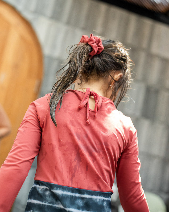 Eine Person mit nassen Haaren, die mit einem Haargummi zu einem Pferdeschwanz gebunden sind, trägt einen roten Laila Mädchen-Badeanzug von Main Design aus umweltfreundlichem Stoff mit LSF 50+ Sonnenschutz und ist von hinten aufgenommen. Sie steht vor einer Holztür und einer strukturierten Steinmauer, was darauf hindeutet, dass sie sich im Freien befindet.