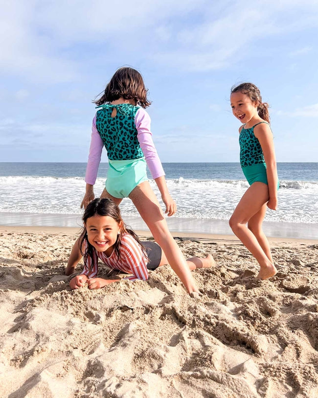 Drei lächelnde Mädchen genießen einen Sandstrand im Laila Mädchen Badeanzug von Main Design aus recycelten Materialien. Zwei stehen, während eines im Sand liegt, während sich unter einem wolkenverhangenen Himmel die Wellen nähern.