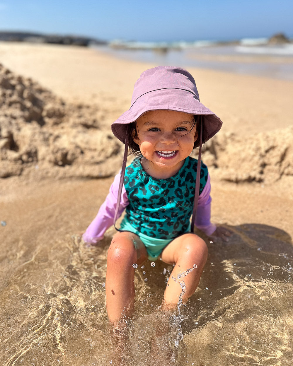 Ein kleines Kind mit einem lilafarbenen Sonnenhut und dem türkisfarbenen Leopardenprint Laila Mädchen Badeanzug von Main Design sitzt lächelnd im flachen Wasser an einem sonnigen Sandstrand.