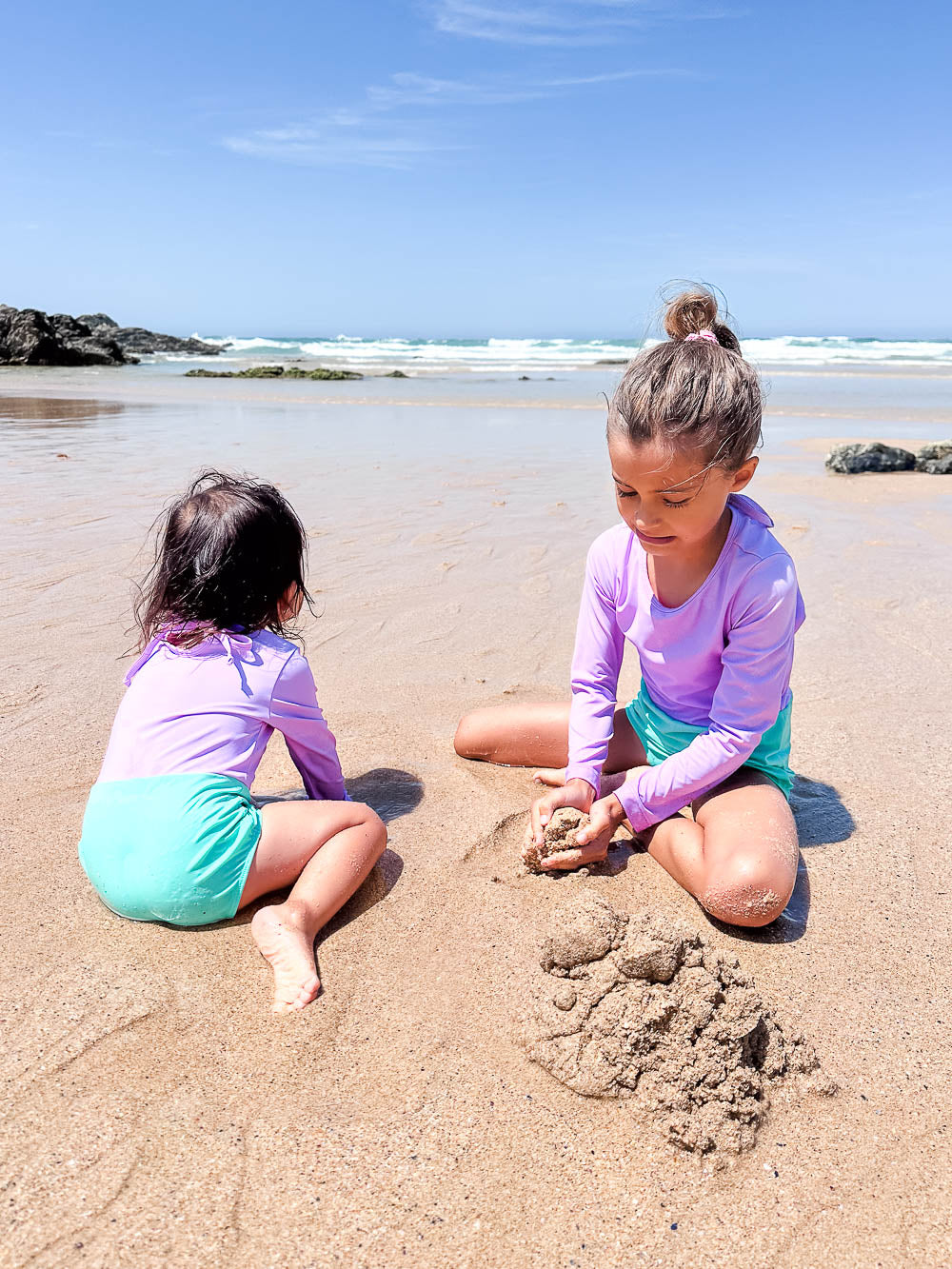 Zwei kleine Kinder im passenden Laila Mädchen Badeanzug von Main Design mit UPF 50+ spielen im Sand an einem sonnigen Strand, mit Wellen und Felsen im Hintergrund.