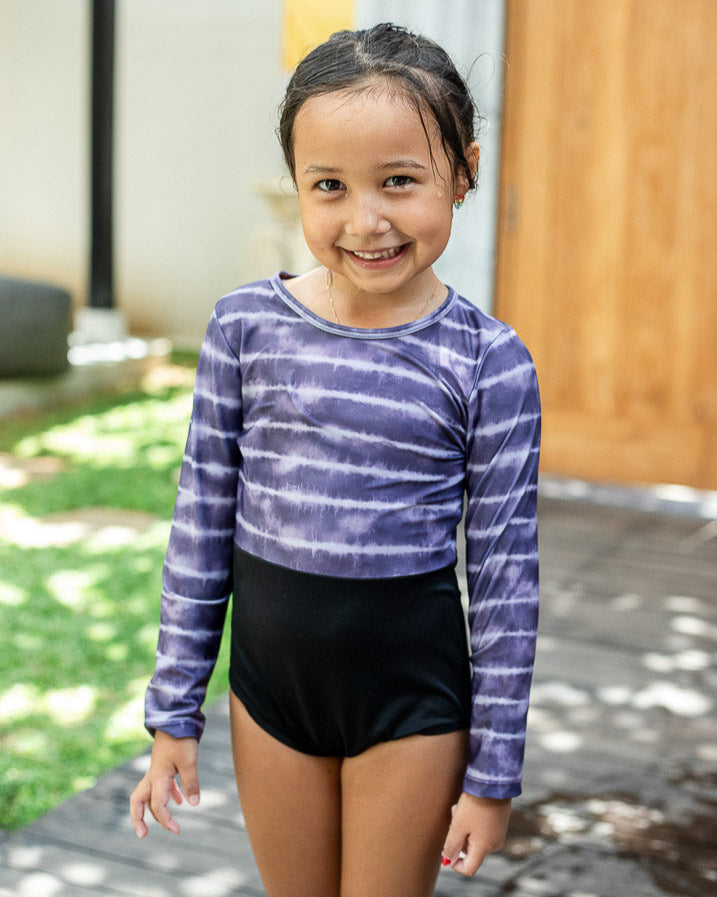 Ein junges Mädchen in einem lilafarbenen Laila Mädchen-Badeanzug mit Batikmuster von Main Design und schwarzen Shorts lächelt in die Kamera. Sie steht draußen auf einer Holzfläche mit viel Grün und einer Holztür im Hintergrund. Das schnell trocknende Material sorgt dafür, dass sie sich den ganzen Tag lang wohl fühlt.