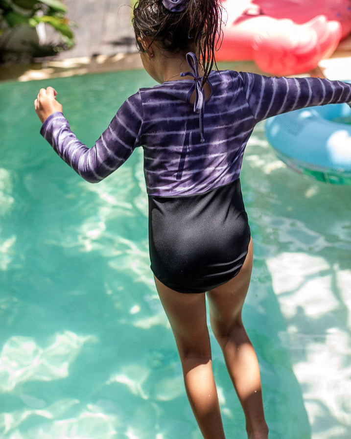 Ein Kind in einem langärmligen, zweifarbigen Badeanzug springt in ein Schwimmbecken. Der Laila Mädchen Badeanzug von Main Design hat oben ein lila-weißes Batikmuster und unten ein einfarbiges Schwarz. Mit einem Haargummi im Haar sind im Hintergrund bunte Schwimmhilfen zu sehen.