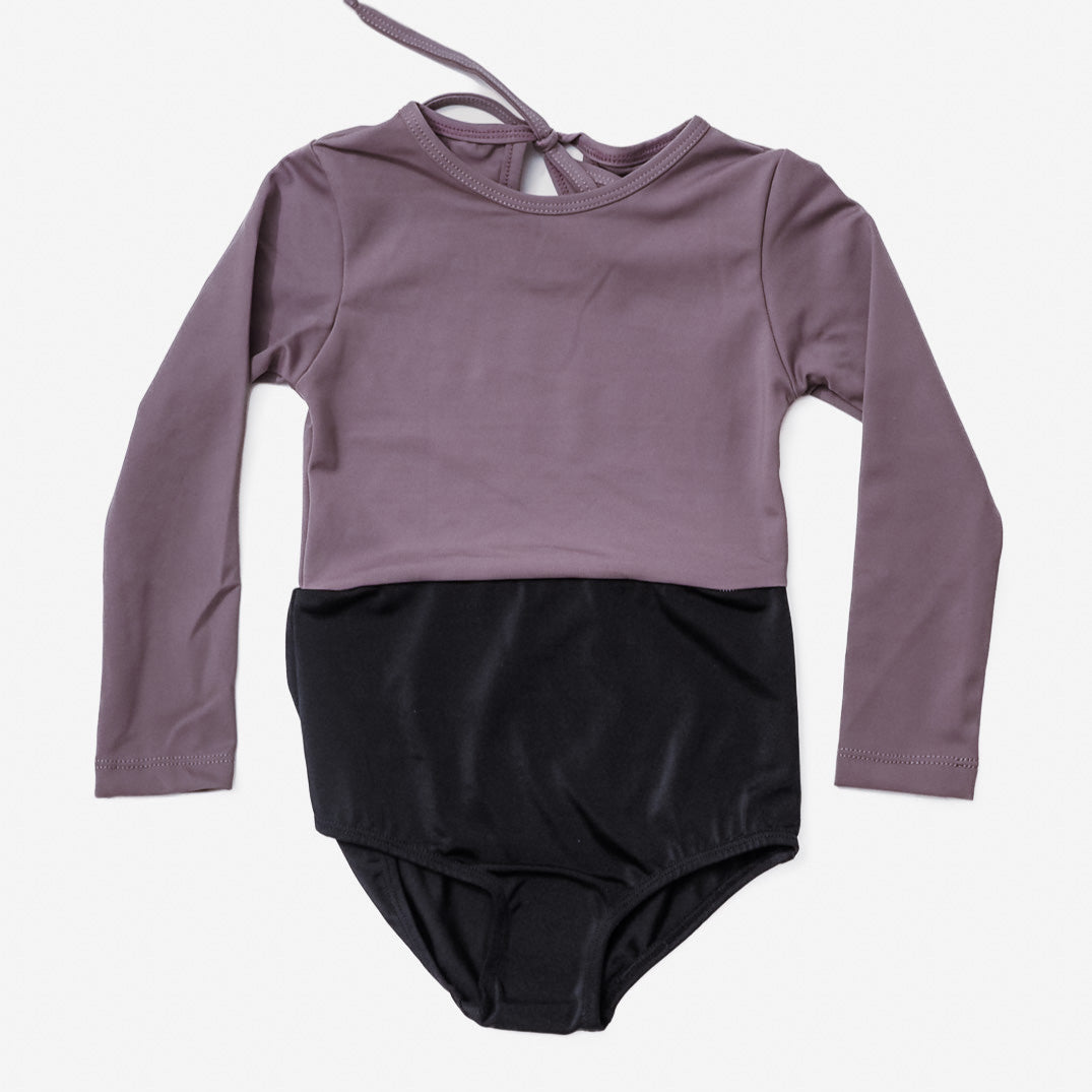 Der Laila Mädchen Badeanzug von Main Design ist ein langarmiger, zweifarbiger Leotard mit einem malvenfarbenen Oberteil mit LSF 50+ Sonnenschutz und einem schwarzen Unterteil. Er verfügt über einen Bindeverschluss auf der Rückseite und ist wunderschön auf weißem Hintergrund dargestellt, ideal für alle, die stilvolle und dennoch schützende Badebekleidung suchen.