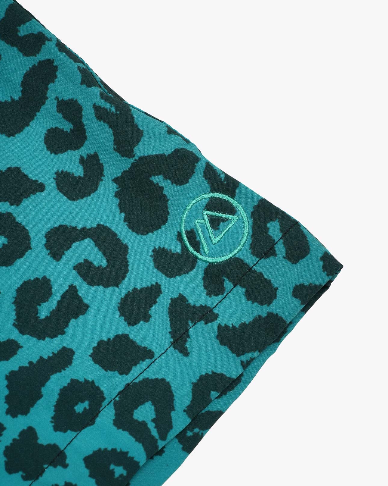 Nahaufnahme des blaugrünen Stoffes mit dunklem Leopardenmuster und 4-Wege-Stretch. Ein kleines, rundes Logo mit einem umrandeten Dreieck befindet sich am Rand, ideal für die Lean Boardshorts für Jungen von Main Design mit UPF 50+ Sonnenschutz.