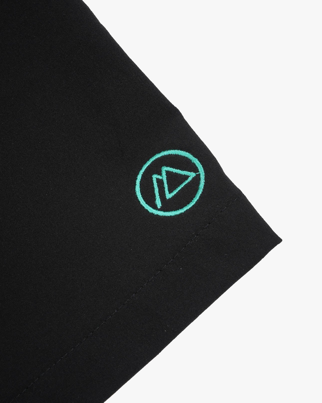 Nahaufnahme des schwarzen 4-Wege-Stretch-Stoffes der Lean Boardshorts für Jungen von Main Design mit einem grün gestickten Logo, das einem stilisierten Dreieck in einem Kreis ähnelt. Sauber vernähte Kanten sorgen für Strapazierfähigkeit und Stil.