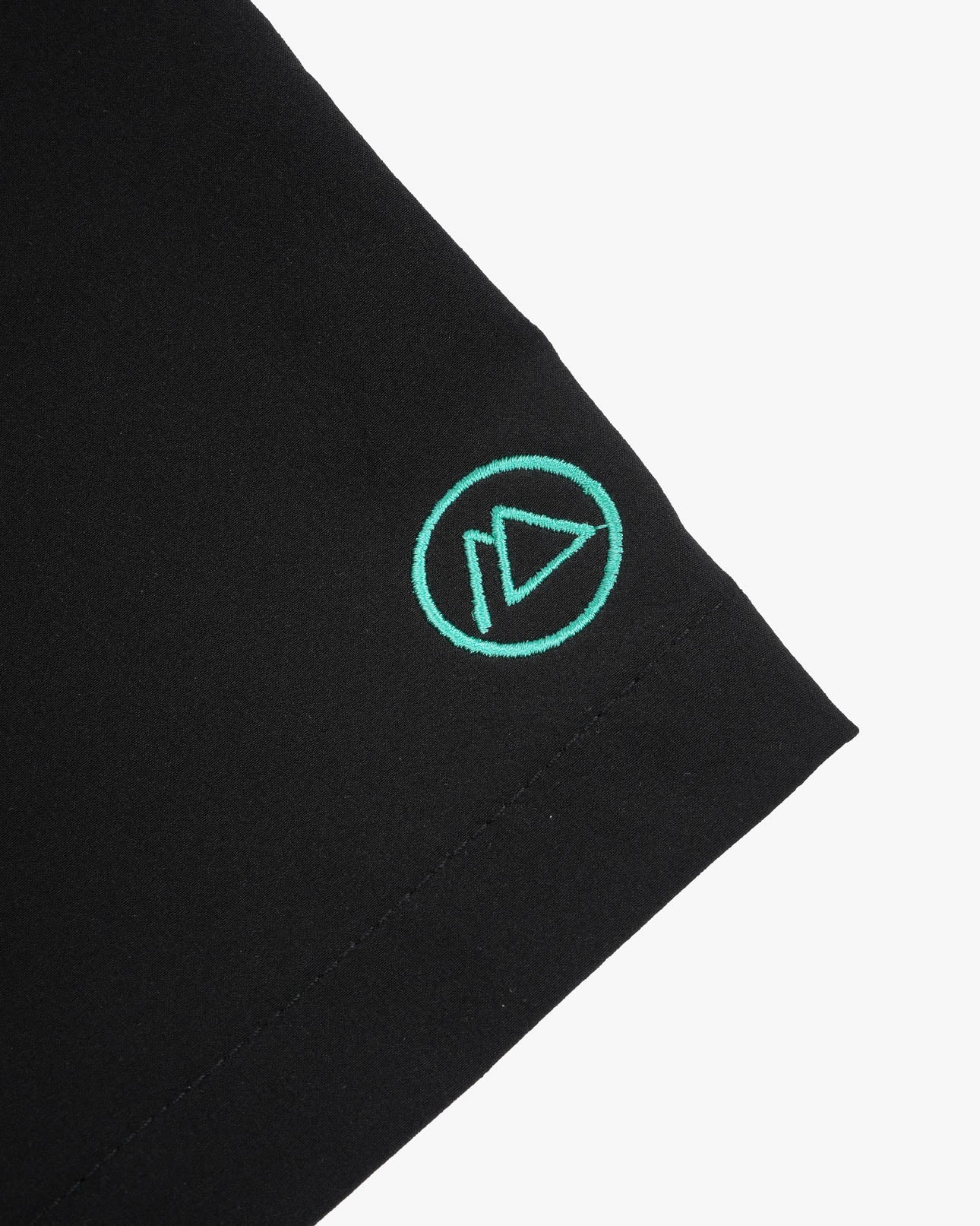 Nahaufnahme des schwarzen 4-Wege-Stretch-Stoffes der Lean Boardshorts für Jungen von Main Design mit einem grün gestickten Logo, das einem stilisierten Dreieck in einem Kreis ähnelt. Sauber vernähte Kanten sorgen für Strapazierfähigkeit und Stil.