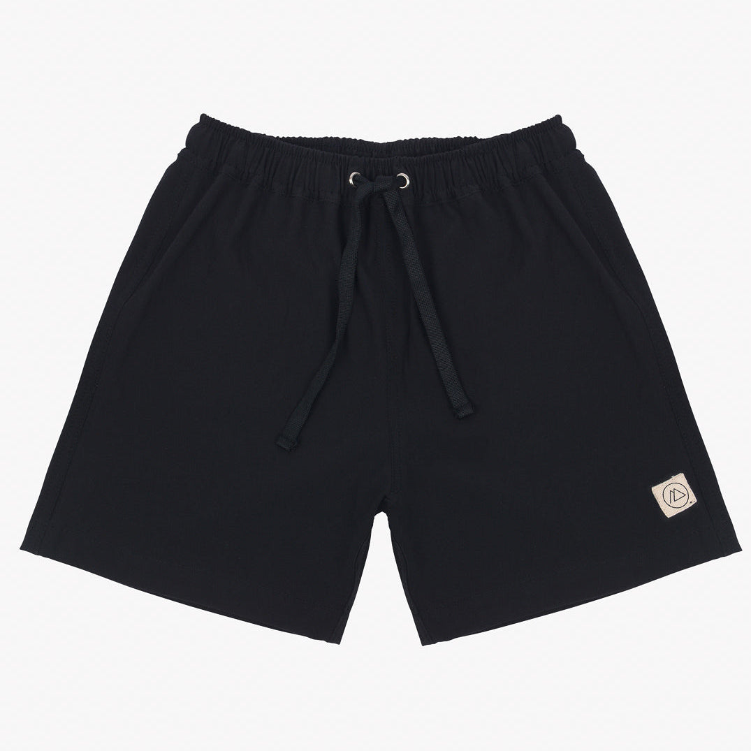 Ein Paar schwarze Main Design Lean Boardshorts für Jungen mit elastischem Bund und schwarzem Kordelzug. Die Shorts haben zwei Seitentaschen, 4-Wege-Stretch-Material, Sonnenschutzfaktor 50+ und einen kleinen rechteckigen beigen Patch mit einem Logo auf der unteren linken Seite.