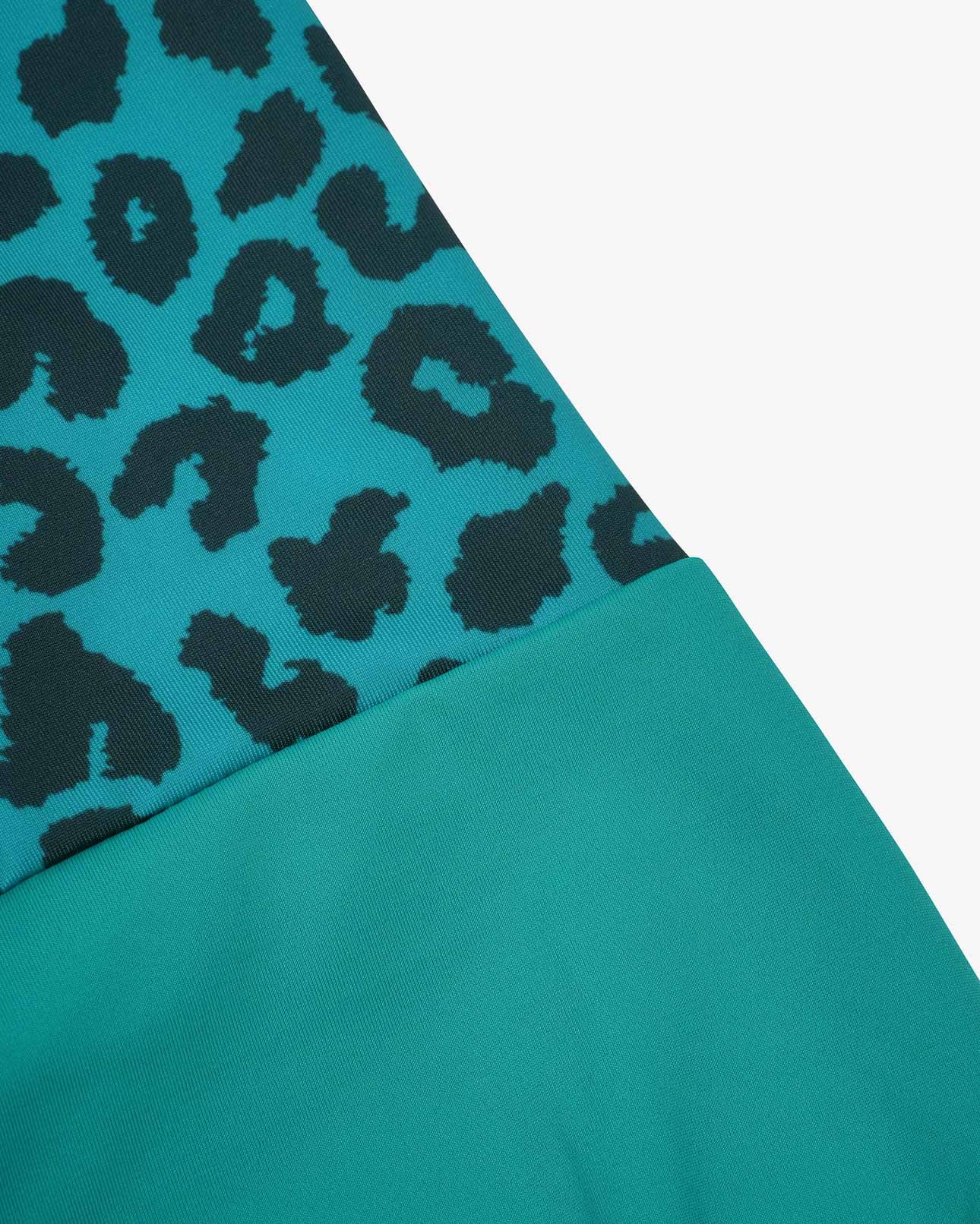 Nahaufnahme eines türkisfarbenen Lena Mädchen-Badeanzugs von Main Design mit Leopardenmuster aus recycelten Materialien. Der Stoff hat eine glatte Textur mit unregelmäßigen schwarzen Formen auf einer Seite und einfarbigem Türkis auf der anderen, wodurch ein stilvoller diagonaler Übergang entsteht.