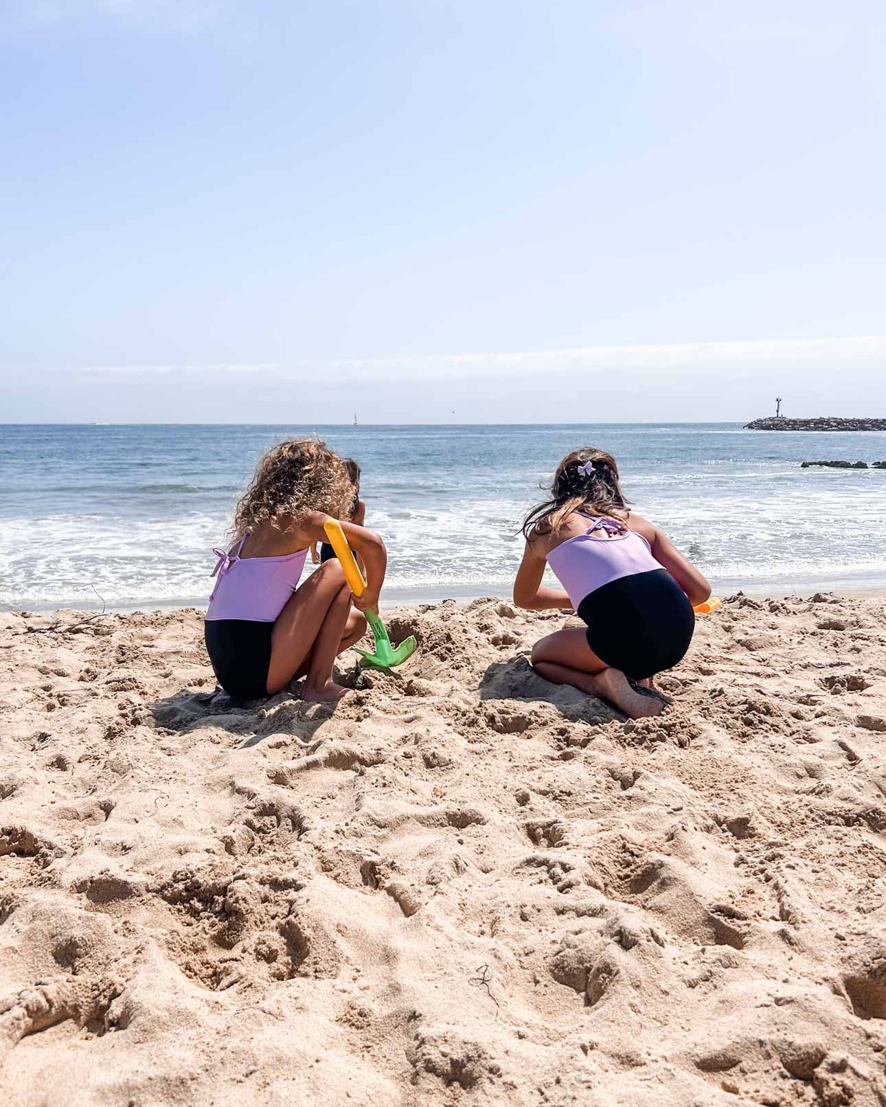 Zwei junge Mädchen im Lena Mädchen Badeanzug von Main Design knien an einem Sandstrand und graben mit bunten Schaufeln. Ihr Blick geht unter klarem Himmel auf das Meer hinaus. Hinter ihnen rollen Wellen heran, und rechts erscheint ein Steg.