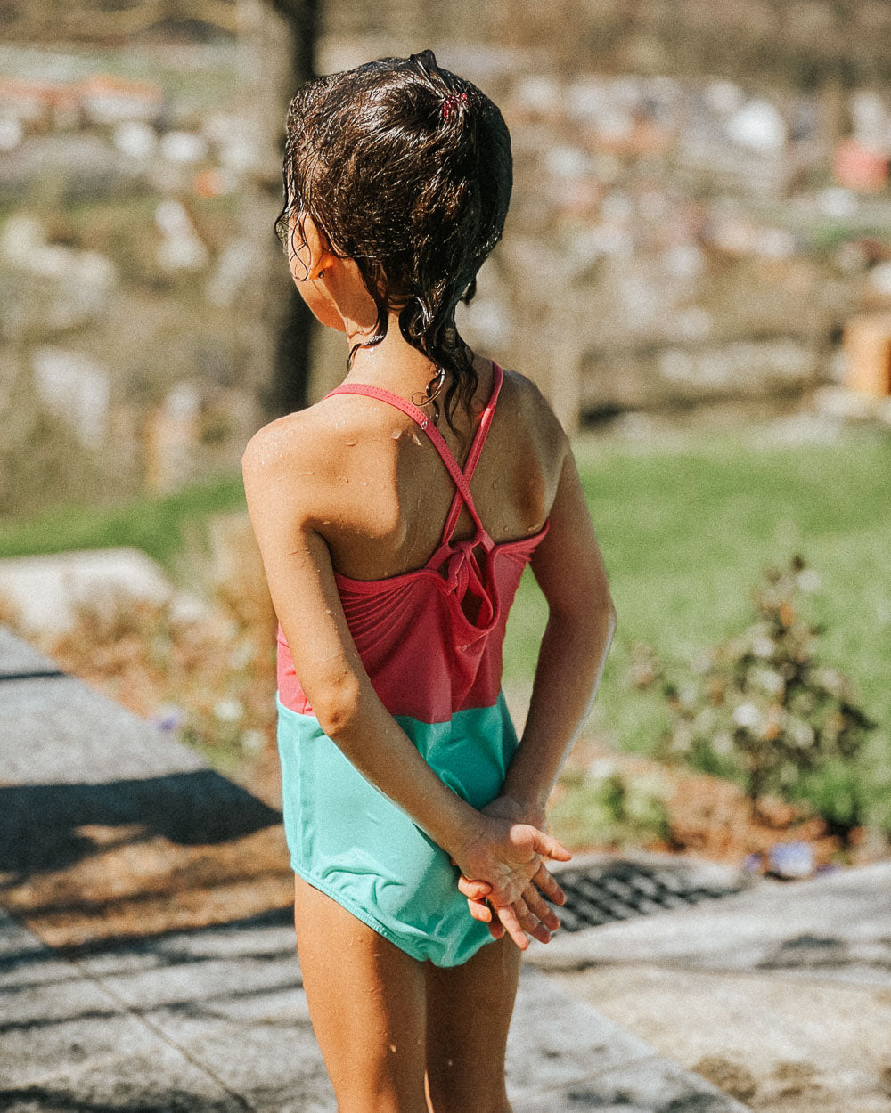 Ein junges Mädchen mit nassen Haaren steht im Freien in einem pink- und blaugrünen Lena Mädchen Badeanzug von Main Design aus recycelten Materialien und blickt mit hinter dem Rücken verschränkten Händen vor einem grasbewachsenen, unscharfen Hintergrund in die Ferne.