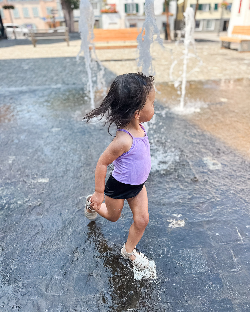 Ein kleines Kind in einem lilafarbenen Lena Mädchen Badeanzug von Main Design und schwarzen Shorts läuft an einem sonnigen Tag fröhlich durch einen Wasserbrunnen auf einem gepflasterten öffentlichen Platz.