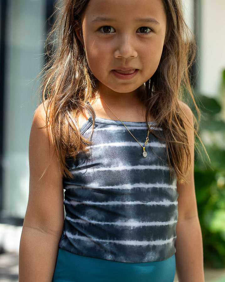 Ein junges Mädchen mit langen braunen Haaren steht im Freien, trägt einen Lena Mädchen Badeanzug von Main Design und eine blaue Hose. Sie trägt eine Halskette mit einem kleinen Anhänger und blickt mit einem leichten Lächeln in die Kamera. Im Hintergrund sind ein Gebäude und Grünflächen zu sehen.