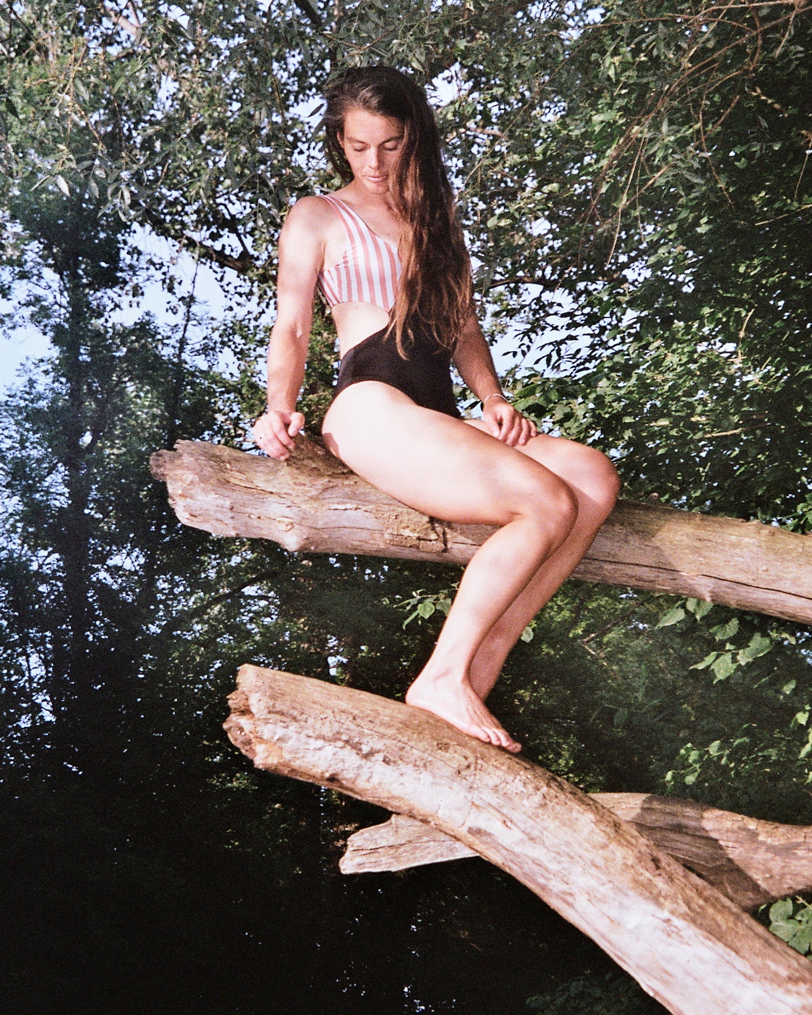 Im Badeanzug Luna von Main Design mit seitlichen Ausschnitten sitzt eine Frau auf einem großen Ast über dem Wasser inmitten von üppigem Grün und natürlichem Licht und blickt entspannt nach unten.