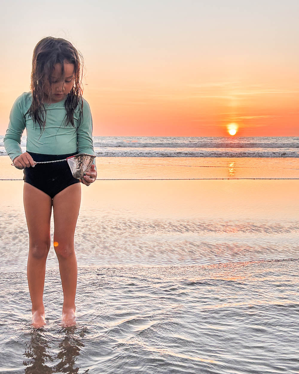 Ein junges Mädchen im Laila Mädchen Badeanzug von Main Design steht bei Sonnenuntergang barfuß im seichten Strandwasser und hält sanft einen Fisch. Der UPF 50+ Schutz und das nachhaltige Design zeigen ihre Sorge um die Natur und den Komfort.