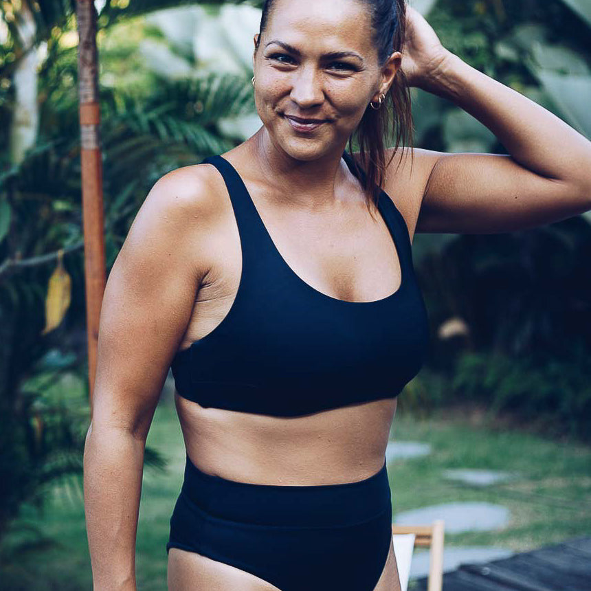 Eine Frau, die die schwarze Alice Bikini Hose von Main Design trägt, lächelt und berührt ihr Haar in einem üppigen grünen Garten, umgeben von tropischen Pflanzen und verschwommenen Outdoor-Möbeln, die eine nachhaltige Sommeratmosphäre schaffen.