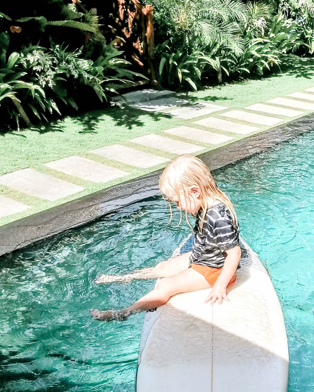 Ein junges blondes Kind sitzt auf einem Surfbrett am Pool, trägt das Main Design Aina Kinder UV Shirt (UPF 50+) und taucht seine Füße ins Wasser, umgeben von üppigen Grünpflanzen und einem Steinweg.