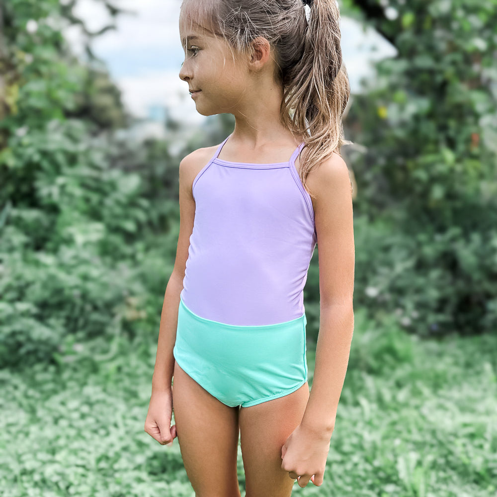 Ein junges Mädchen mit hellbraunem Haar in einem Pferdeschwanz steht auf grünem Gras, trägt den Lena Mädchen Badeanzug von Main Design in Lavendel und Mintgrün und blickt nach links, mit Bäumen und Pflanzen im Hintergrund.