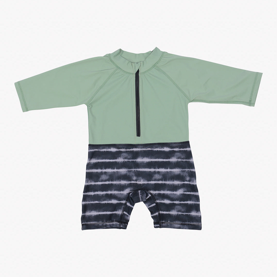 Der Malia Baby Badeanzug von Main Design ist ein langärmliger Baby-Strampler mit einem grünen Oberteil mit einem schwarzen Reißverschluss in der Mitte und einem schwarz-weißen Batik-Unterteil. Der Strampler hat einen hohen Halsausschnitt, ist für bequemes Tragen konzipiert und bietet LSF 50+ für zusätzlichen Sonnenschutz.