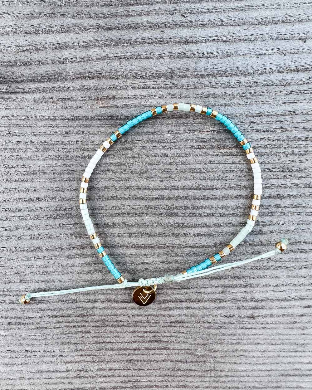 Das Mama und Mini Armband Wave von Main Design ist ein handgefertigtes Schmuckstück mit türkis, weißen und goldenen Perlen sowie einem kleinen runden Checkmark-Charm - nachhaltig präsentiert auf grauer Struktur.