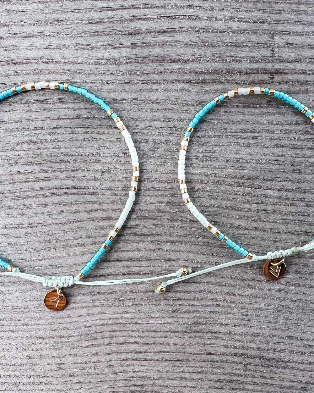 Zwei Main Design Mama und Mini Armband Wave Armbänder liegen nebeneinander auf einer grauen Oberfläche, mit blauen, weißen und goldenen Perlen und einem braunen runden Anhänger für nachhaltigen Schmuck.