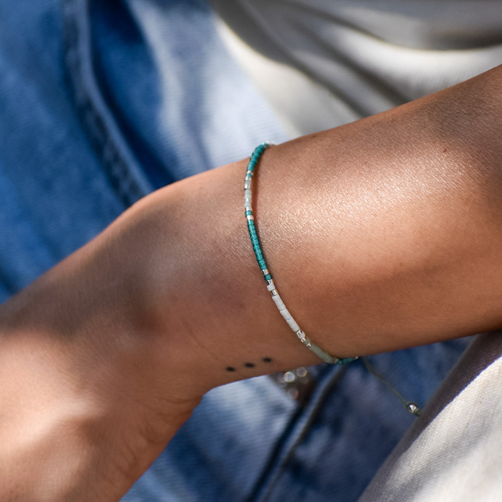 Eine Nahaufnahme eines Handgelenks mit einem Drei-Punkte-Tattoo, das das Mama und Mini Armband Wave von Main Design trägt. Die Person trägt eine hellblaue Denim-Jeans und ein helles Top, das nachhaltigen Schmuck hervorhebt.