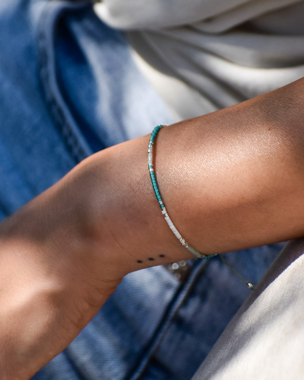 Eine Nahaufnahme eines Handgelenks mit einem Drei-Punkte-Tattoo, das das Mama und Mini Armband Wave von Main Design trägt. Die Person trägt eine hellblaue Denim-Jeans und ein helles Top, das nachhaltigen Schmuck hervorhebt.