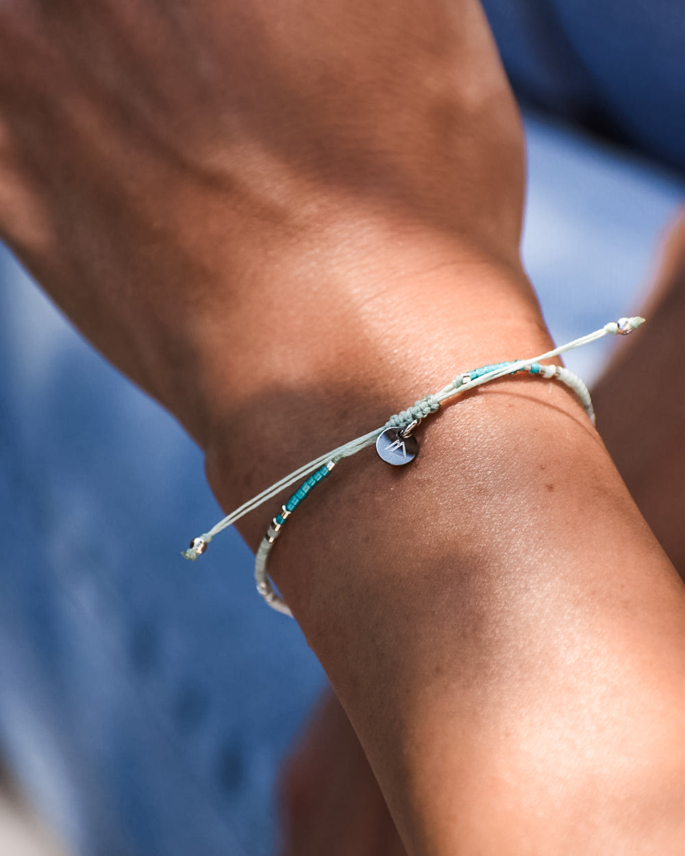 Nahaufnahme eines Handgelenks, das zwei zarte Kordelarmbänder von Main Design trägt: das Mama und Mini Armband Wave in Hellblau mit Perlen und einem kleinen runden Anhänger sowie ein weißes Armband, das mit hellblauer Jeanskleidung kombiniert wird.