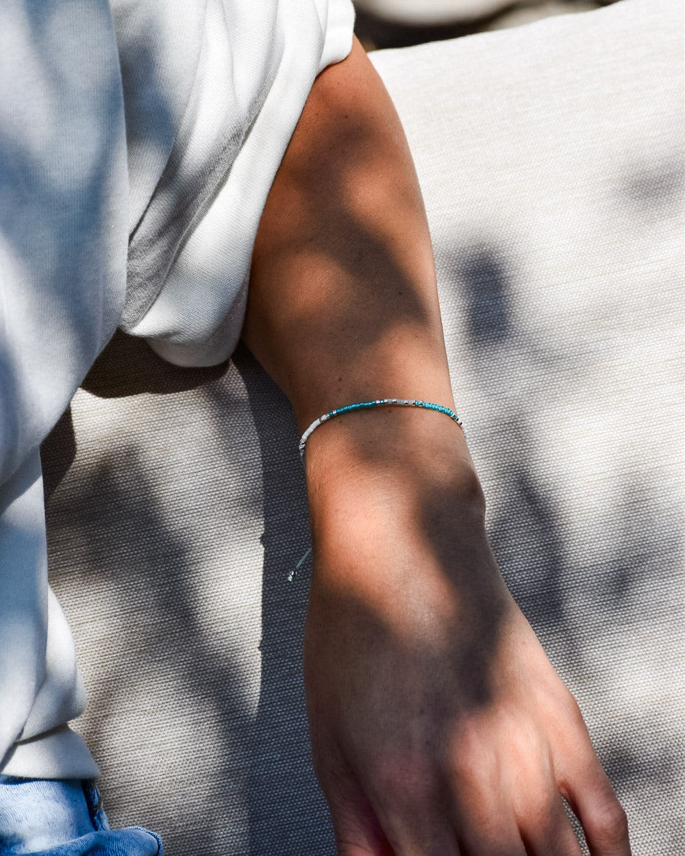 Eine Person trägt das filigrane Mama und Mini Armband Wave von Main Design in Türkis und legt ihren Arm auf hellen Stoff, während Sonnenlicht und Schatten sanft über die Haut spielen - ein Beispiel für nachhaltigen Schmuck.