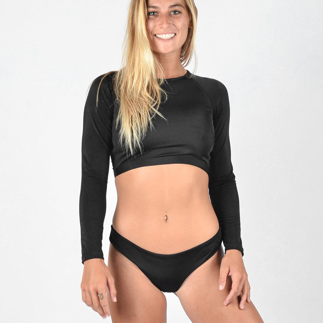 Eine Frau steht lächelnd in die Kamera. Sie hat langes blondes Haar und trägt ein schwarzes, langärmliges Lycra-Crop-Top von Maria Langarm mit einem schwarzen Bikinihöschen von Main Design. Der Hintergrund ist schlicht weiß.