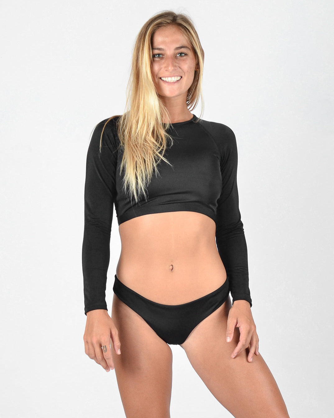 Eine Frau steht lächelnd in die Kamera. Sie hat langes blondes Haar und trägt ein schwarzes, langärmliges Lycra-Crop-Top von Maria Langarm mit einem schwarzen Bikinihöschen von Main Design. Der Hintergrund ist schlicht weiß.