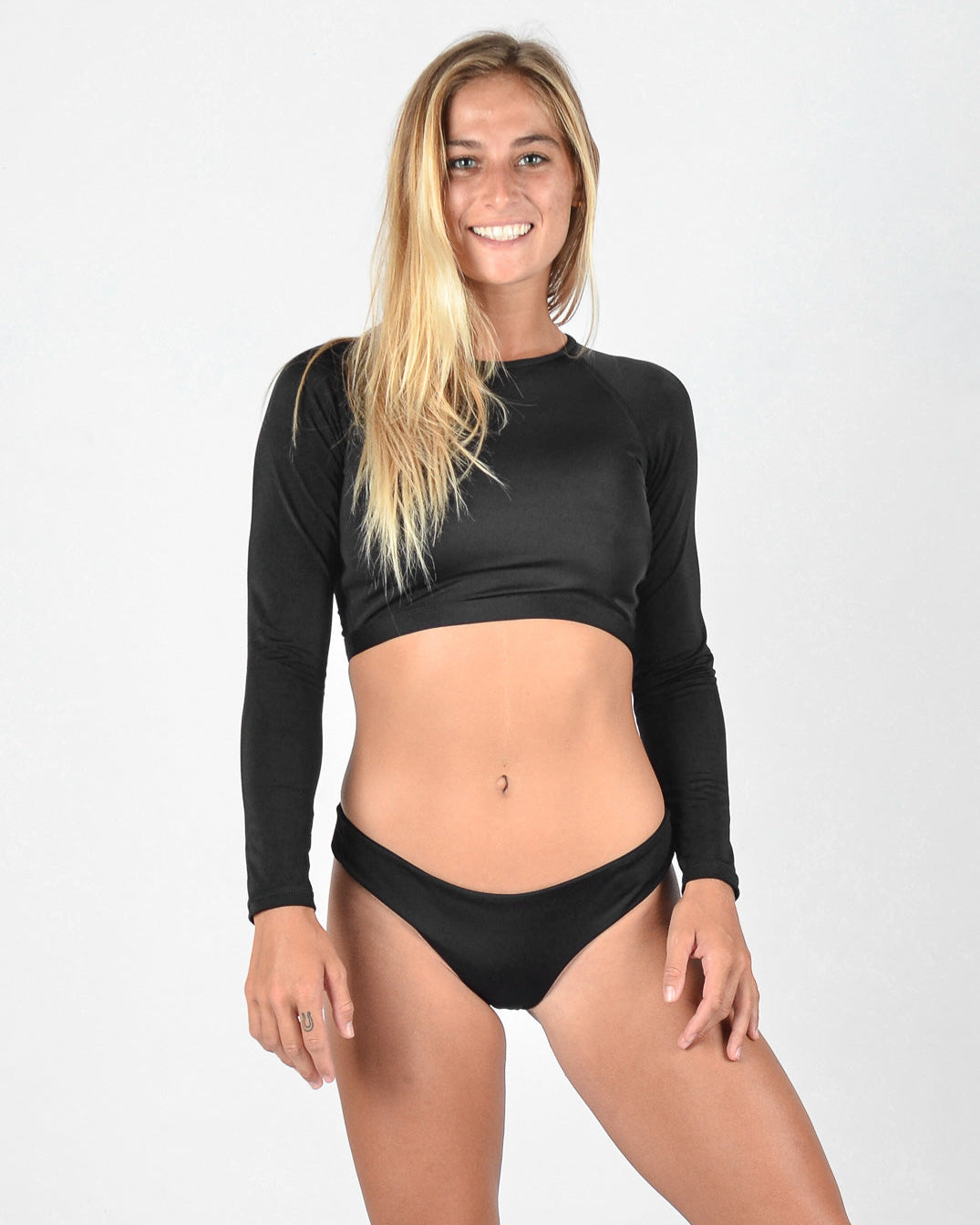 Eine Frau steht lächelnd in die Kamera. Sie hat langes blondes Haar und trägt ein schwarzes, langärmliges Lycra-Crop-Top von Maria Langarm mit einem schwarzen Bikinihöschen von Main Design. Der Hintergrund ist schlicht weiß.