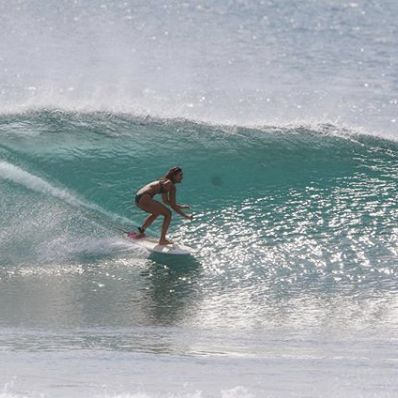 Eine Person im Badeanzug surft auf einer klaren, türkisfarbenen Welle, tief geduckt auf ihrem Surfbrett, während das Sonnenlicht auf dem Wasser funkelt.
