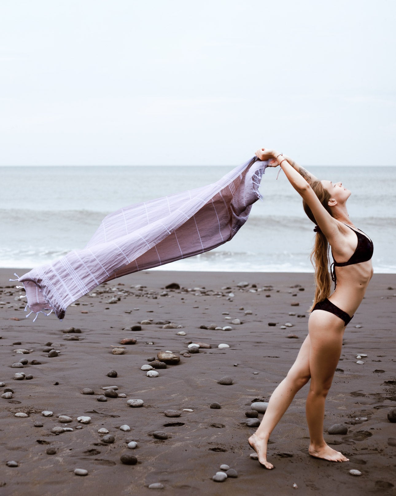 Eine Frau in einem schwarzen Bikini steht barfuß an einem felsigen Strand und streckt ihre Arme nach hinten, während sie das Main Design Maxi Strandtuch De La Mer in der Hand hält, mit dem Meer und dem bewölkten Himmel im Hintergrund.