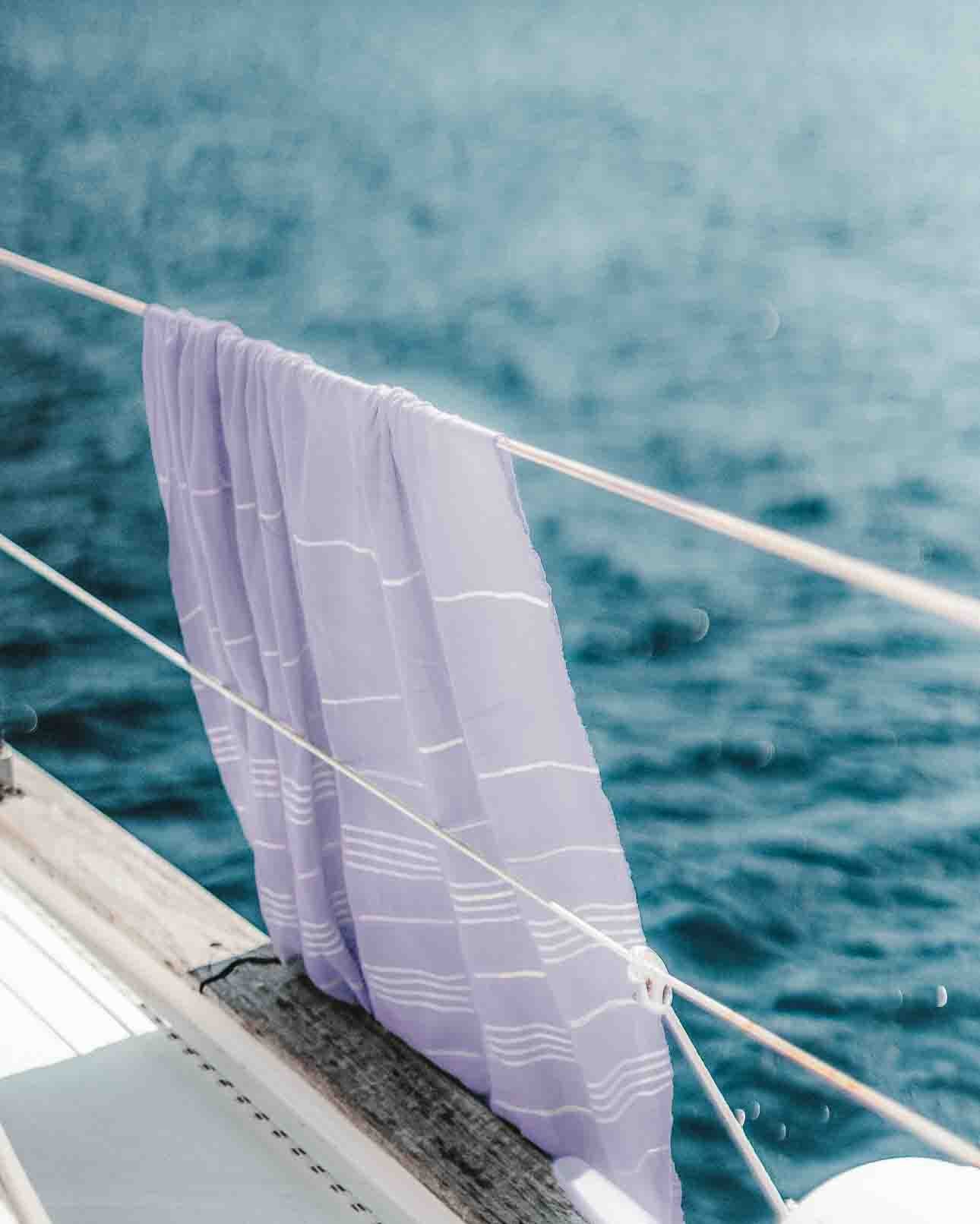 Ein Maxi Strandtuch De La Mer von Main Design mit lavendelfarbenen Streifen aus hochwertiger Baumwolle hängt über dem Bootsreling, im Hintergrund ruhiges blaues Wasser.