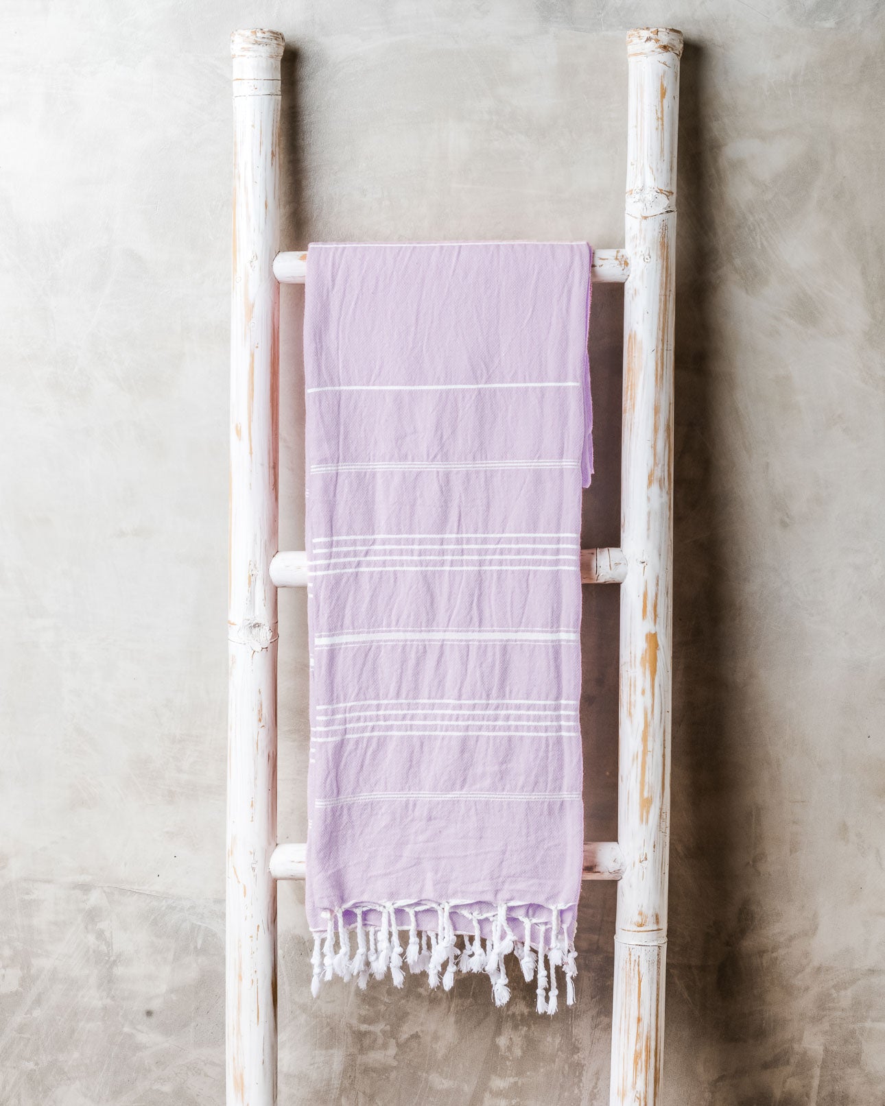 Ein hellviolettes Maxi Strandtuch De La Mer von Main Design aus hochwertiger Baumwolle mit weißen Streifen und Fransen liegt über einer weißen Holzleiter vor einer grauen Wand.