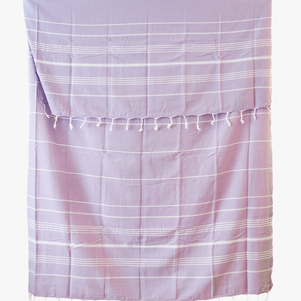 Das Maxi Strandtuch De La Mer von Main Design ist ein lavendelfarbenes Strandtuch aus 100% Baumwolle mit dünnen weißen Streifen und Quasten am oberen und unteren Rand. Es ist OEKO-TEX-zertifiziert und liegt flach auf einem weißen Hintergrund.