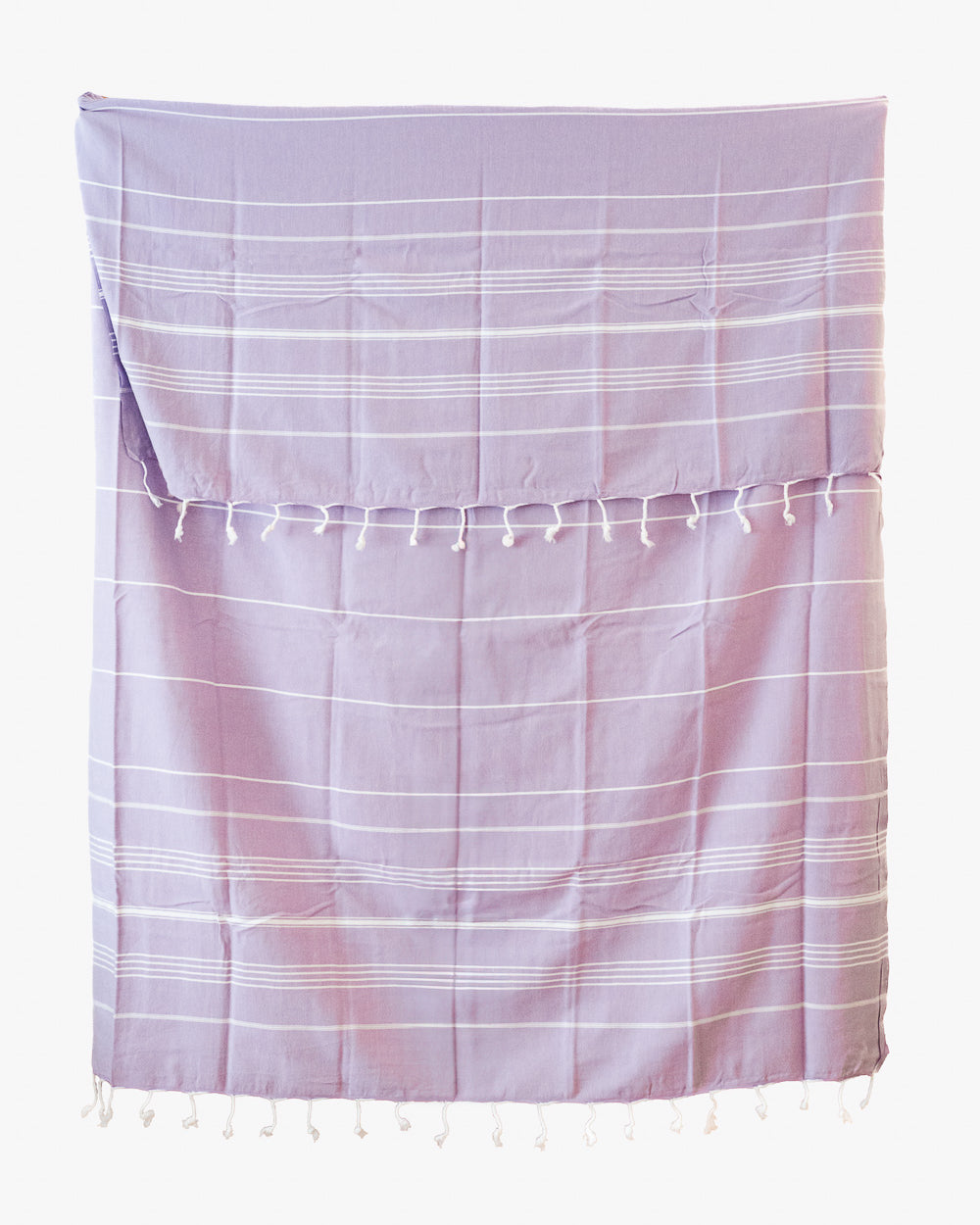 Das Maxi Strandtuch De La Mer von Main Design ist ein lavendelfarbenes Strandtuch aus 100% Baumwolle mit dünnen weißen Streifen und Quasten am oberen und unteren Rand. Es ist OEKO-TEX-zertifiziert und liegt flach auf einem weißen Hintergrund.